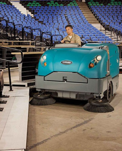 Operator using a Tennant S30 ride-on sweeper to clean concrete floors inside a large indoor stadium with tiered seating.