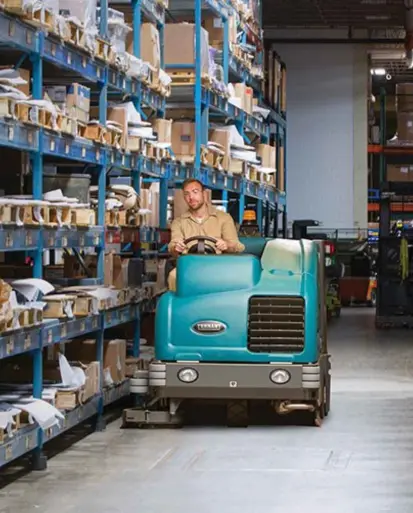 Warehouse worker operating a Tennant ride-on floor scrubber through narrow aisles filled with industrial shelving and cardboard packaging